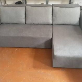 Szary narożnik tapicerowany z trzema poduszkami o jaśniejszym odcieniu, ustawiony na pomarańczowej podłodze. Widoczny fragment półki w tle.