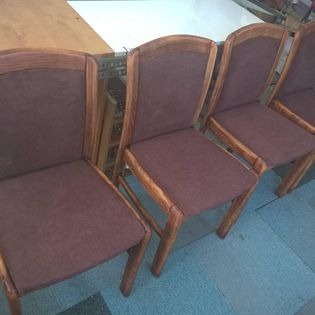 Cztery krzesła tapicerowane w brązowej tkaninie, z drewnianymi ramami w ciepłym odcieniu brązu, ustawione rzędem na podłodze, widoczne w dziennym świetle.