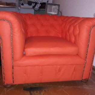 Pomarańczowy fotel typu chesterfield z pikowanym oparciem i siedziskiem, ozdobiony nitami, sfotografowany w słabym oświetleniu na tle nieuporządkowanego pomieszczenia.