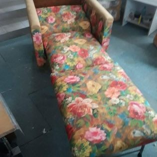 Rozłożony fotel tapicerowany wzorem w duże, kolorowe kwiaty na beżowym tle, widoczne elementy metalowej konstrukcji, niedoskonałe oświetlenie pomieszczenia.