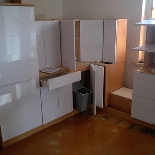 Montaż białej zabudowy kuchennej z drewnianymi elementami, widoczna wysunięta szuflada z dokumentami i kosz na śmieci w przestrzeni szafki pod blatem.