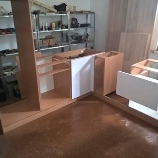 Fragmenty mebli kuchennych w trakcie montażu, widoczne niedokończone szafki z jasnego drewna i białe fronty, na podłodze płyty, w tle metalowy regał z narzędziami i materiałami.
