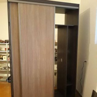 Szafa z przesuwnymi drzwiami w odcieniach brązu i beżu, z otwartymi półkami po prawej stronie, widoczna na tle jasnej ściany i regału z różnymi przedmiotami.