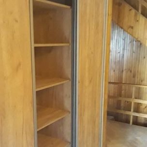 Otwarta szafa wnękowa z jasnego drewna z przesuwnymi drzwiami, widok na wnętrze z półkami, w tle drewniana konstrukcja antresoli pod skośnym dachem.