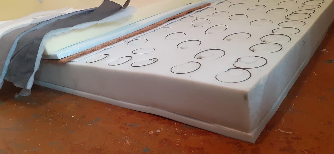 Warstwy materiałów do tapicerowania: pianka z zaznaczonymi okręgami, włókno kokosowe, jasna pianka, szara tkanina i biała wata.