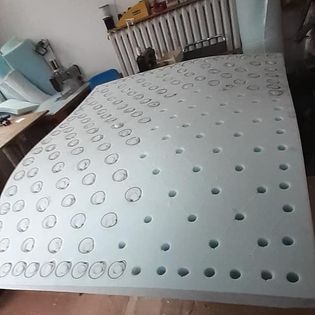 Arkusz jasnoniebieskiej pianki tapicerskiej z wyciętymi okrągłymi otworami, leżący na stole w warsztacie, widoczne narzędzia i maszyna do szycia w tle.