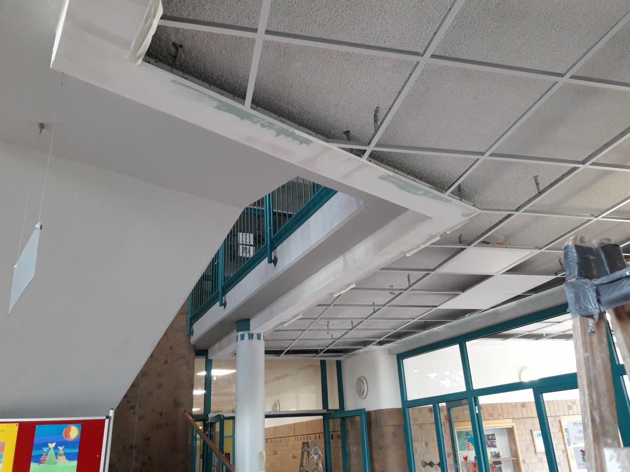 Sufit podwieszany kasetonowy w trakcie montażu w budynku użyteczności publicznej, widoczne elementy konstrukcyjne, szpachlowane łączenia na ścianie i podciągu, fragment balustrady schodów.