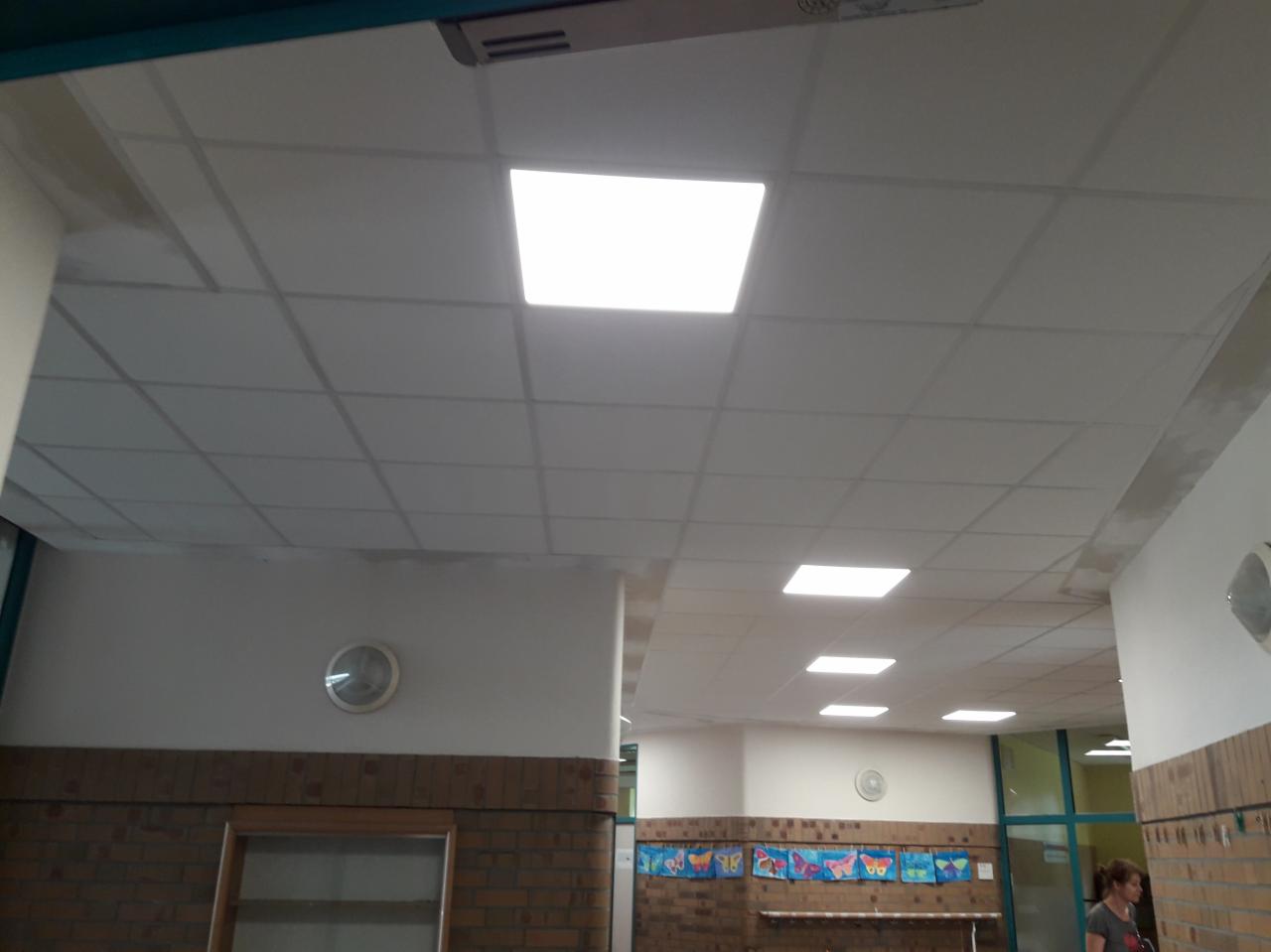 Kasetonowy sufit podwieszany z kwadratowymi panelami oświetleniowymi w korytarzu budynku, widoczne ślady po pracach wykończeniowych na łączeniach paneli.