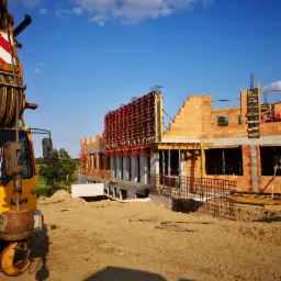 Wznoszenie dwukondygnacyjnego domu z czerwonej cegły silikatowej, z widocznym szalunkiem na ścianach i żółtym dźwigiem w pierwszym planie, na placu budowy pokrytym piaskiem.