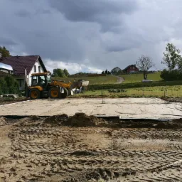 Żółta koparko-ładowarka przygotowuje teren pod fundamenty na działce z widokiem na domy na wzgórzu, pochmurne niebo.