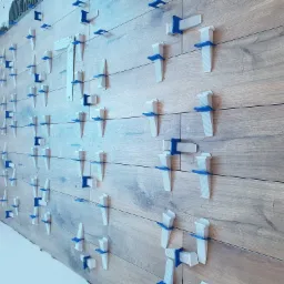 Ściana z drewnopodobnych płytek, w trakcie montażu, z użyciem białych i niebieskich klinów poziomujących, widoczne ślady zaprawy na krawędziach.