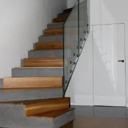 Schody drewniane z betonowymi podstopnicami i szklaną balustradą, widoczne również szklane drzwi w tle.