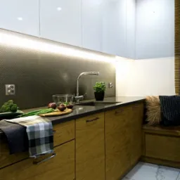 Nowoczesna kuchnia z drewnianymi szafkami dolnymi i białymi szafkami górnymi, oświetleniem LED pod szafkami, na blacie warzywa przygotowane do gotowania, oraz siedzisko z poduszkami w rogu kuchni.