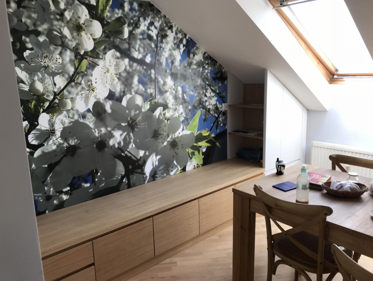 Adaptacja poddasza: zabudowa z szafkami pod skosem ściany z fototapetą z motywem kwitnących gałęzi, drewniany stół z krzesłami i okno dachowe