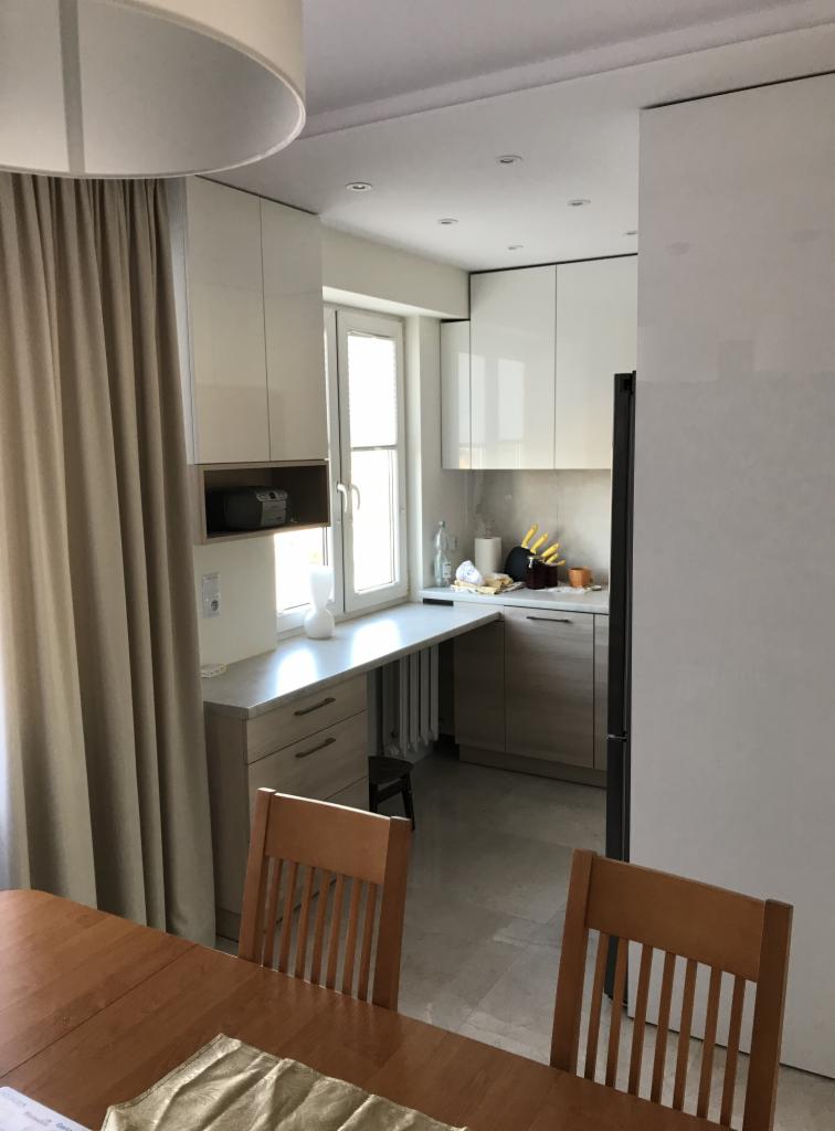 Jasna kuchnia z białymi szafkami, blatem roboczym pod oknem i drewnianymi frontami szuflad. Widoczne krzesła i stół w jadalni.