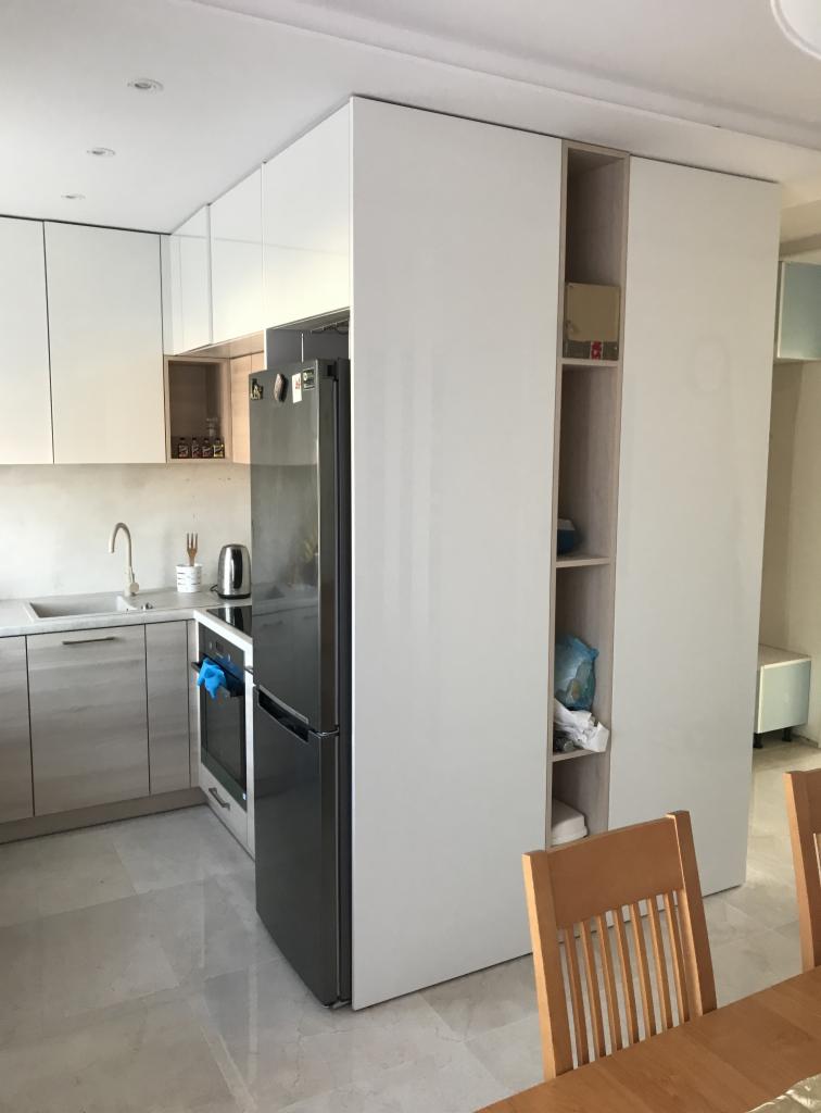 Nowoczesna kuchnia z zabudową meblową w odcieniach bieli i szarości, z widoczną lodówką i szafą wnękową z otwartymi półkami, oświetlona naturalnym światłem.