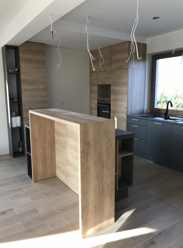 Wnętrze kuchni w trakcie montażu, widoczne wiszące kable od oświetlenia, drewnopodobna zabudowa oraz szare szafki z blatem pod oknem, na podłodze płytki imitujące drewno.