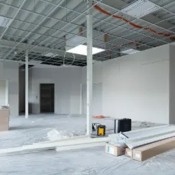 Wnętrze w trakcie remontu: surowe ściany z płyt gipsowo-kartonowych, metalowa konstrukcja sufitu podwieszanego, podłoga zabezpieczona folią, widoczne narzędzia i materiały budowlane, w tym profile...