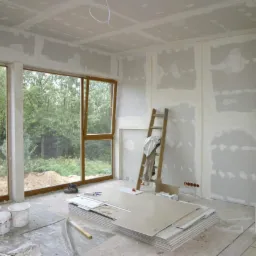 Wnętrze w trakcie prac wykończeniowych: ściany i sufit pokryte gładzią gipsową, widoczne płyty kartonowo-gipsowe, drabina oparta o ścianę, duże okna z widokiem na zieleń, przygotowane farby...