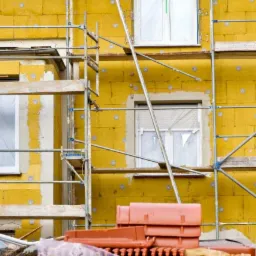 Żółta elewacja budynku w trakcie ocieplania, widoczne rusztowania i okna zabezpieczone folią, fragmenty starego tynku i cegieł, elementy instalacyjne.