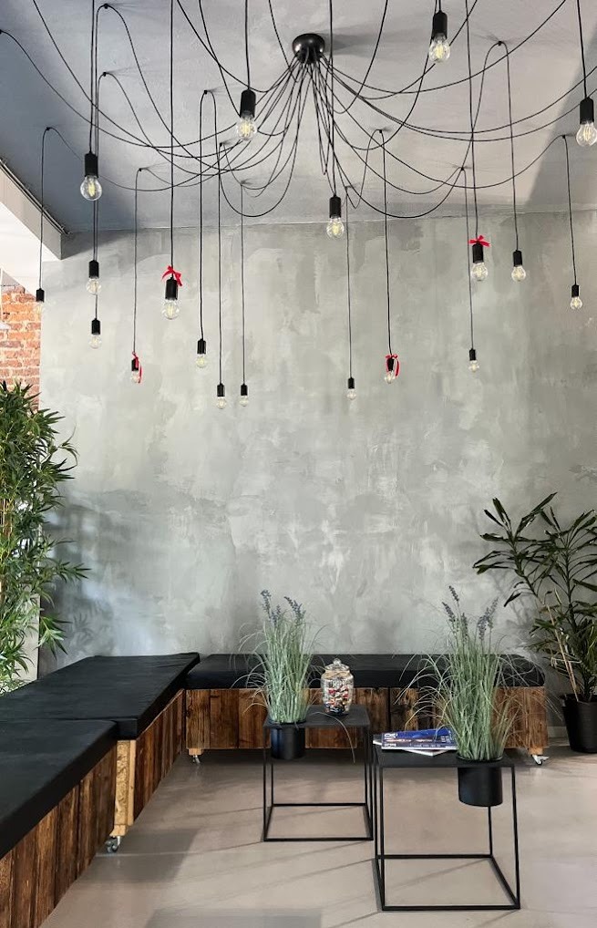 Nowoczesne wnętrze salonu z betonową ścianą, wiszącymi żarówkami z czerwonymi wstążkami, drewnianymi ławkami z czarnymi poduszkami i roślinami w donicach.