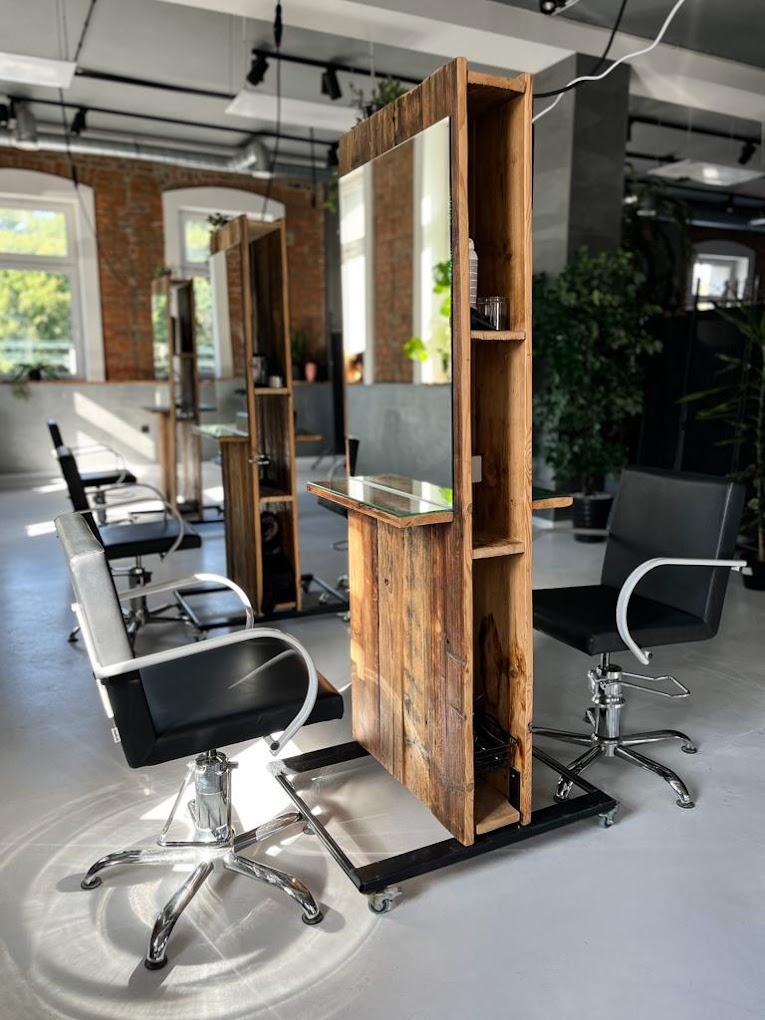 Wnętrze salonu z trzema stanowiskami do stylizacji włosów, każde wyposażone w lustro w drewnianej obudowie na kółkach i czarne krzesło z chromowaną podstawą, w tle ceglana ściana i duże okna.