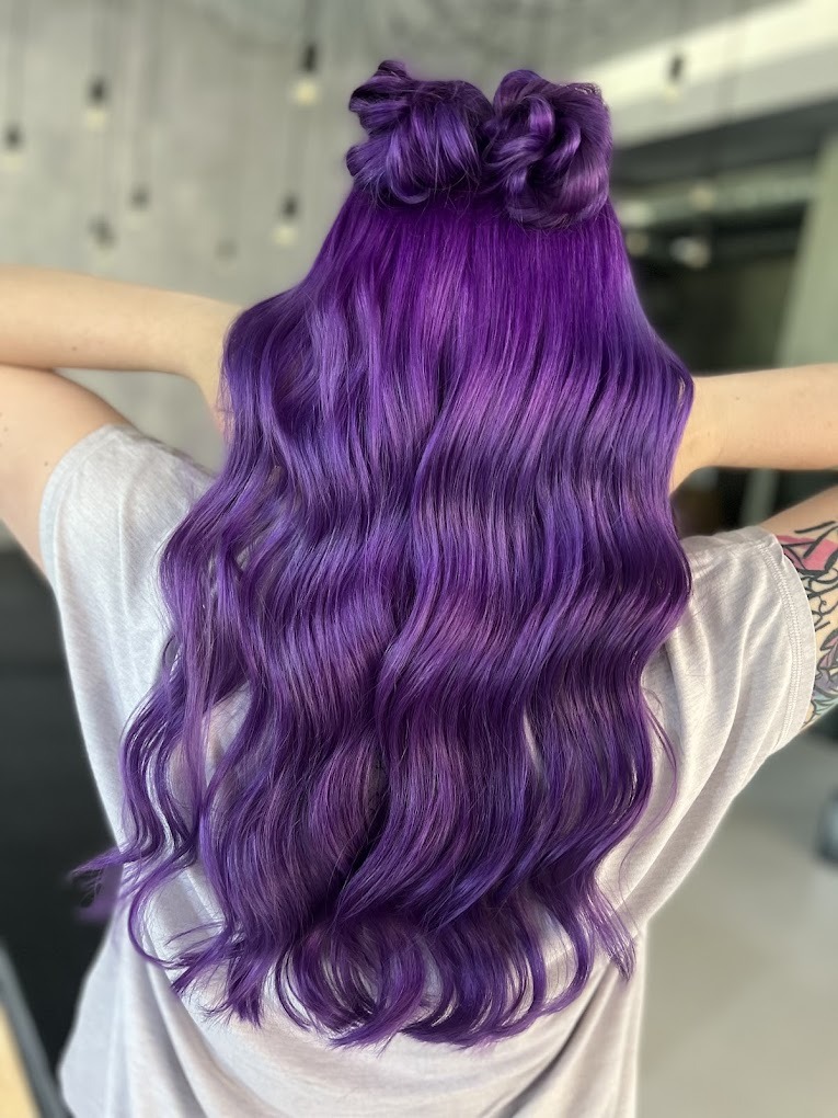 Długie, falowane włosy w odcieniach fioletu z dwoma małymi kokami na czubku głowy, widoczne od tyłu, z tatuażem na ramieniu.