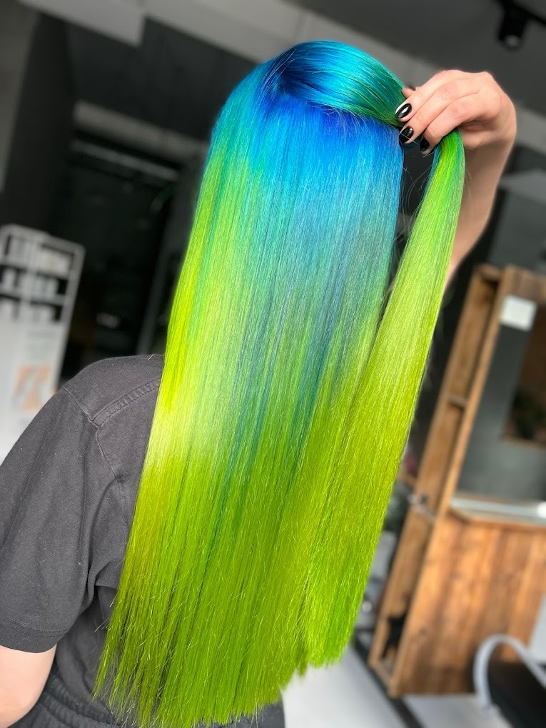 Długie, proste włosy z koloryzacją ombre: od intensywnego błękitu u nasady do jaskrawej limonki na końcach, trzymane w górze dłonią z czarnym manicure.