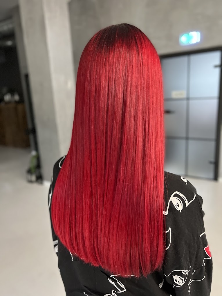 Intensywnie czerwone, proste włosy z ciemniejszym odrostem, widok z tyłu, na tle minimalistycznego wnętrza z szarą ścianą i szklanymi drzwiami.