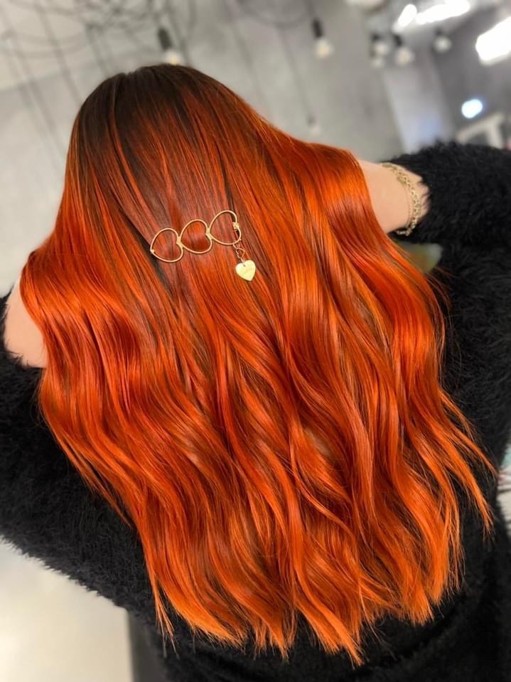 Bujne, lśniące włosy w odcieniach miedzi i rudości, z ciemniejszym odrostem, upięte złotą spinką z motywem trzech kółek i zawieszką w kształcie serca.