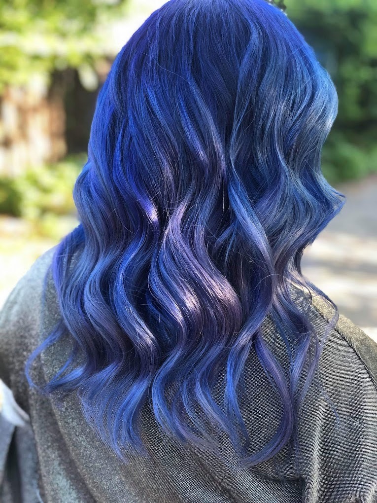 Tył głowy z pofalowanymi włosami w odcieniach intensywnego błękitu i fioletu, delikatnie rozjaśnionymi, na tle rozmytej zieleni.