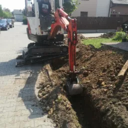 Operator w białej mini koparce Kubota z pomarańczowym ramieniem kopie wąski rów w ziemi przy chodniku z kostki, w tle dom jednorodzinny i zaparkowane samochody.