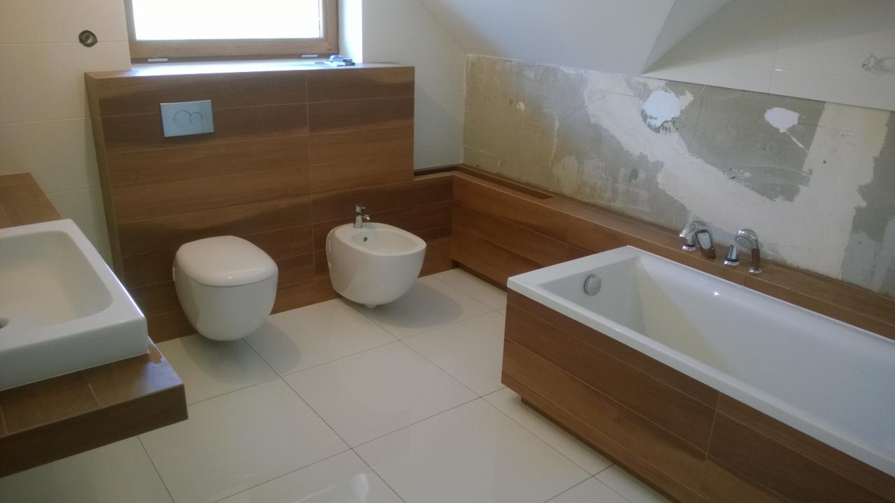 Wnętrze łazienki w trakcie prac wykończeniowych: biała armatura (umywalka, toaleta, bidet, wanna) na tle drewnopodobnych płytek i częściowo wykończonych ścian.