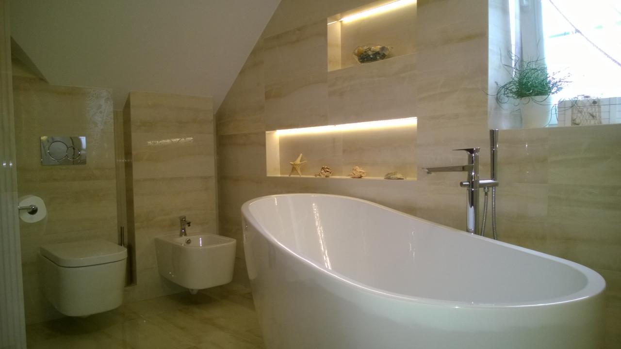 Nowoczesna łazienka z wolnostojącą wanną, podwieszaną toaletą i bidetem, wykończona płytkami imitującymi marmur, z oświetlonymi niszami dekoracyjnymi z muszlami i rozgwiazdą.