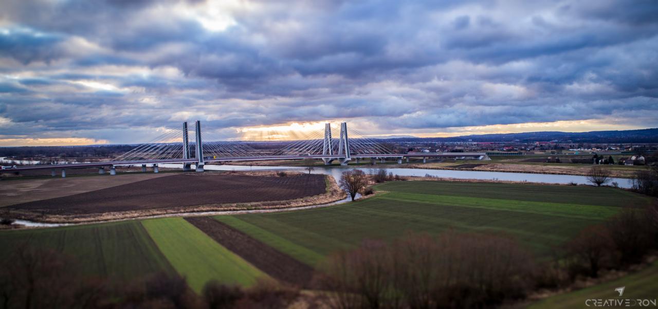 Pejzaż z nowoczesnym mostem wiszącym nad rzeką, pole w paski i zachmurzone niebo z promieniami słońca w tle.