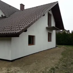 Wykończony dom jednorodzinny z brązowym dachem z dachówki ceramicznej, otynkowaną białą elewacją i drewnianą stolarką okienną. Widoczny fragment ogrodu z niskimi tujami. Nierówny teren wokół budynku.
