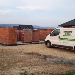 Wczesny etap budowy domu z czerwonej cegły, widoczne fundamenty, częściowo wzniesione ściany, materiały budowlane na paletach oraz firmowy bus 'Smart Dom Kompleksowo' na żwirowym podłożu, w tle...