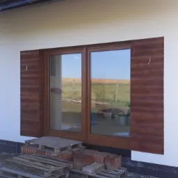 Fragment elewacji domu z dużym oknem przesuwnym, obłożonym dekoracyjną okładziną drewnopodobną, widoczne palety i cegły, sugerujące trwające prace wykończeniowe.
