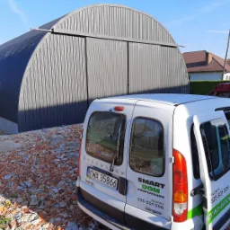 Szara, metalowa hala łukowa z przesuwnymi drzwiami, widoczna zza białego samochodu dostawczego firmy budowlanej na tle nieba.