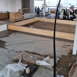 Marfab-Bud Monika Noga - Wnętrze w trakcie budowy, widoczny drewniany szkielet konstrukcji podłogi, materiały budowlane i narzędzia na betonowej posadzce, dwóch pracowników w tle, surowy betonowy strop z widoczną instalacją.