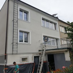 Marfab-Bud Monika Noga - Dwu-kondygnacyjny dom po renowacji elewacji, z jasnoszarą fasadą, białymi obramowaniami okien, z metalową drabiną przymocowaną do ściany, z widocznym fragmentem pracującego mężczyzny na dole...