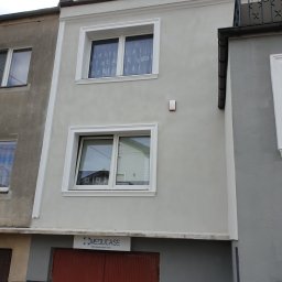 Marfab-Bud Monika Noga - Odnowiona fasada dwupiętrowego domu z nowymi oknami z białymi ramami i dekoracyjnymi listwami, kontrastująca z sąsiednią, starszą elewacją z widocznymi śladami zużycia.