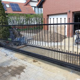Marfab-Bud Monika Noga - Czarna, metalowa brama przesuwna z kulkami na szczytach pionowych prętów, zamontowana przed domem z czerwoną cegłą i panelami słonecznymi na dachu, widoczny fragment kostki brukowej i przygotowany...