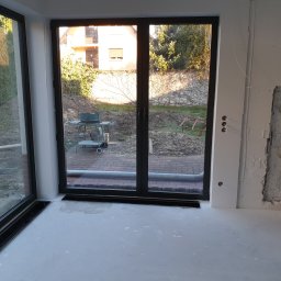 Marfab-Bud Monika Noga - Duże okno przesuwne z czarnymi ramami w nowym domu z widokiem na ogród, ściany w trakcie wykańczania, widoczne instalacje elektryczne.