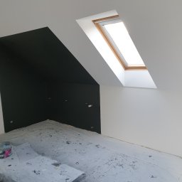 Marfab-Bud Monika Noga - Pokój na poddaszu po malowaniu, ściany białe i ciemnografitowe, okno dachowe z drewnianą ramą, podłoga zabezpieczona folią malarską, widoczny pojemnik po farbie.
