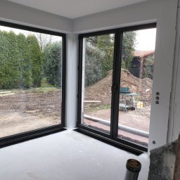 Marfab-Bud Monika Noga - Montaż dużych, przesuwnych okien z czarnymi ramami w Ostroszowicach, widok na ogród z tujami i stertą ziemi, wnętrze w trakcie wykończenia.