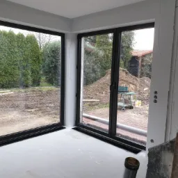 Montaż dużych, przesuwnych okien z czarnymi ramami w Ostroszowicach, widok na ogród z tujami i stertą ziemi, wnętrze w trakcie wykończenia.