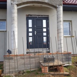 Marfab-Bud Monika Noga - Montaż nowych, grafitowych drzwi wejściowych z szybami w domu jednorodzinnym, widoczne prace murarskie i zbrojenie wokół wejścia, kolumny owinięte taśmą zabezpieczającą.