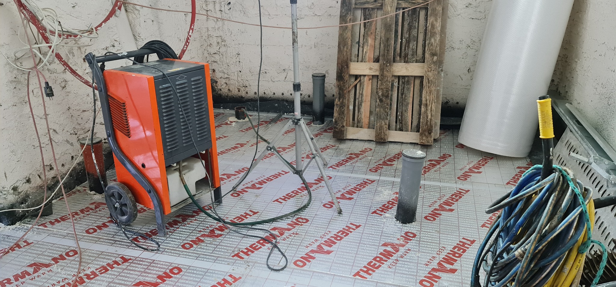 Osuszacz budowlany na kółkach w trakcie pracy, widoczne rury wentylacyjne, podłoże wyłożone folią termoizolacyjną z nadrukiem, paleta drewniana i lampa na statywie w tle.