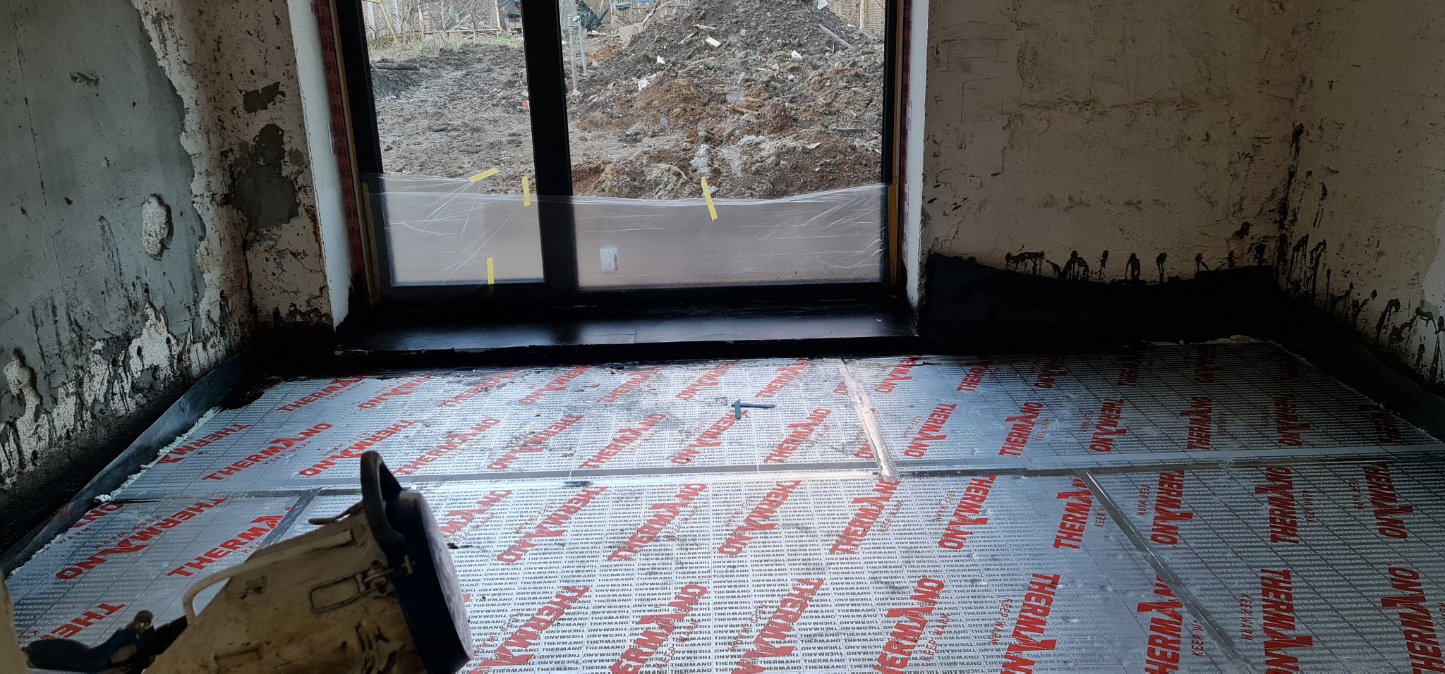 Przygotowanie podłoża pod izolację termiczną podłogi w pomieszczeniu z oknem, widoczne płyty izolacyjne Thermano z czerwonym nadrukiem, surowe ściany z widocznymi śladami po remoncie i narzędzie...